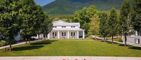 A large white building with a symmetrical facade and columns, surrounded by a well-manicured lawn and trees, with a mountain in the background.
