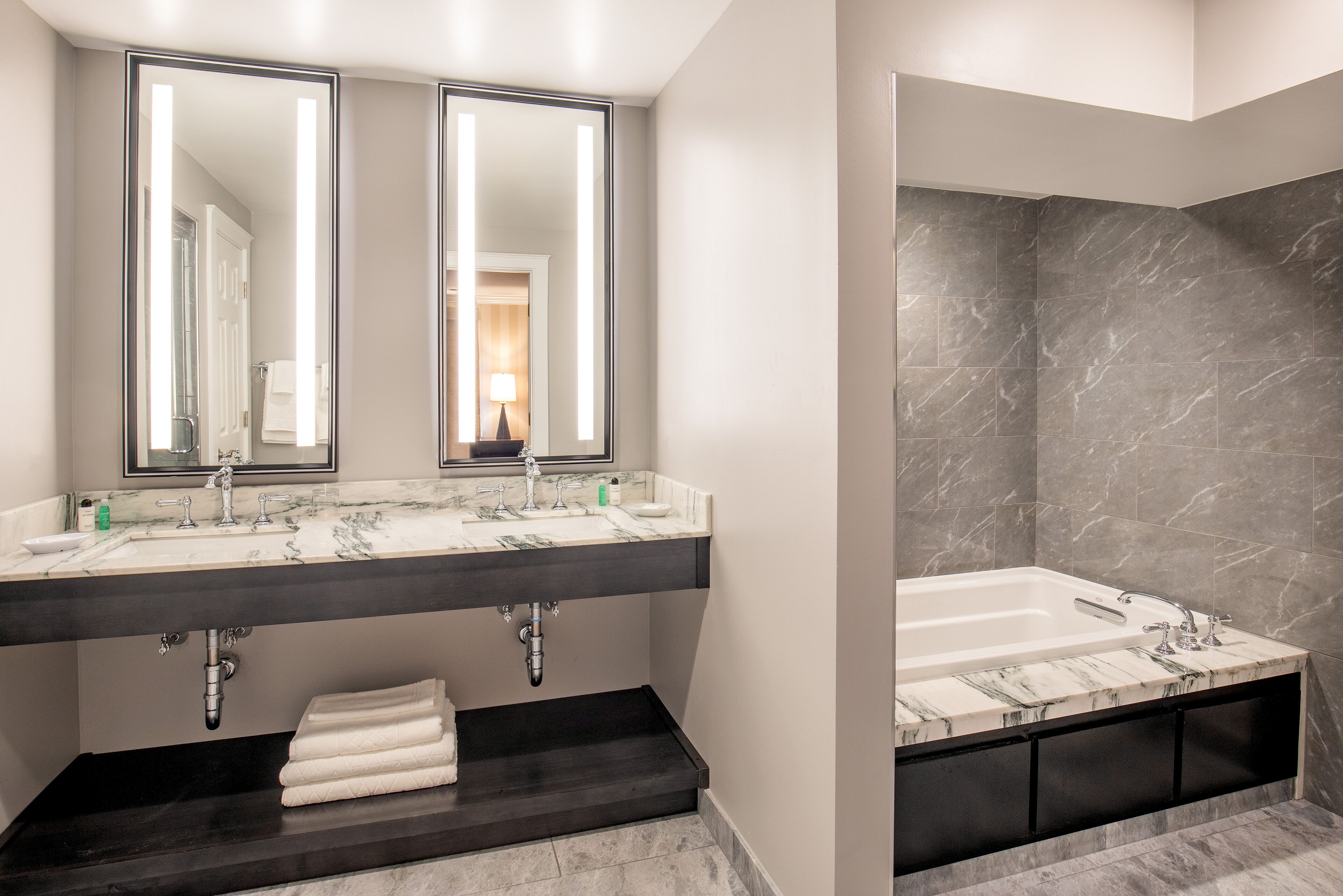 A modern bathroom with a dual sink vanity, a large mirror, a bathtub, and marble flooring.