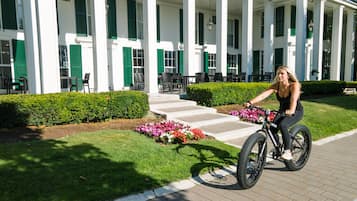 A woman riding a bicycle on a paved path in front of a building with white columns and green shutters.