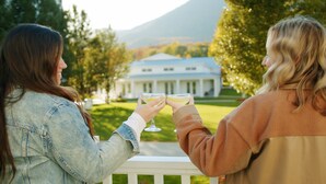 Two people toasting with drinks on a balcony overlooking a large building with columns.