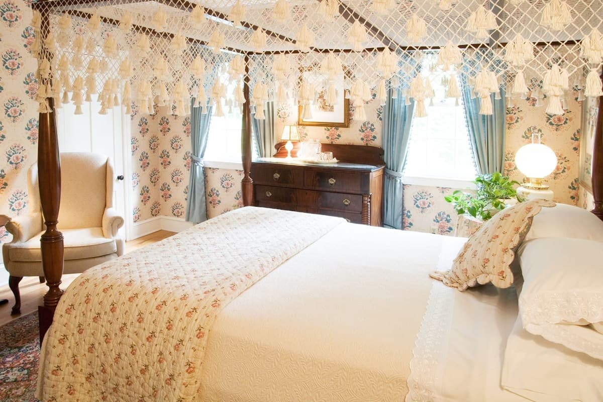 A bedroom with a canopy bed, floral wallpaper, a wooden dresser, and a chair.