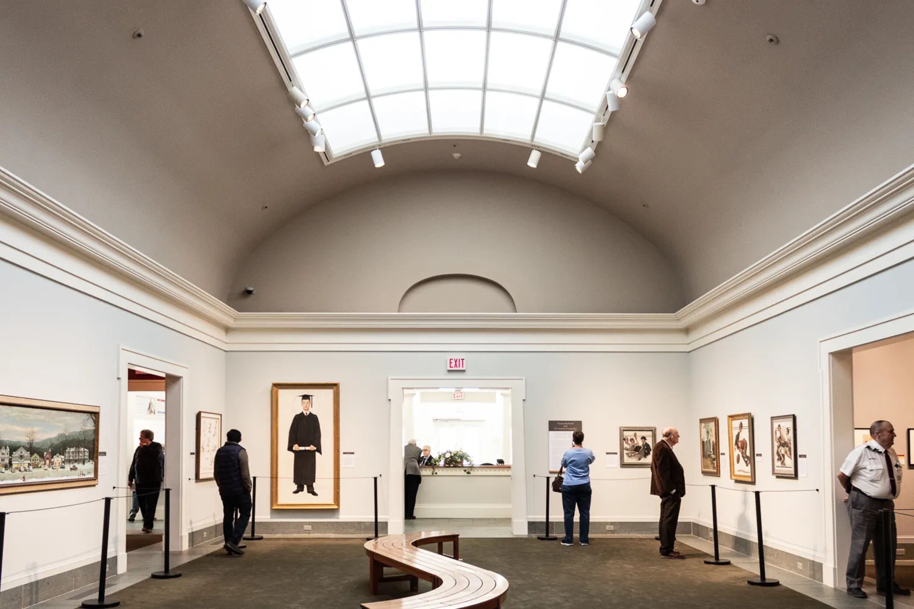 Una galería de arte con techo alto y tragaluces, que exhibe obras enmarcadas y una salida central.