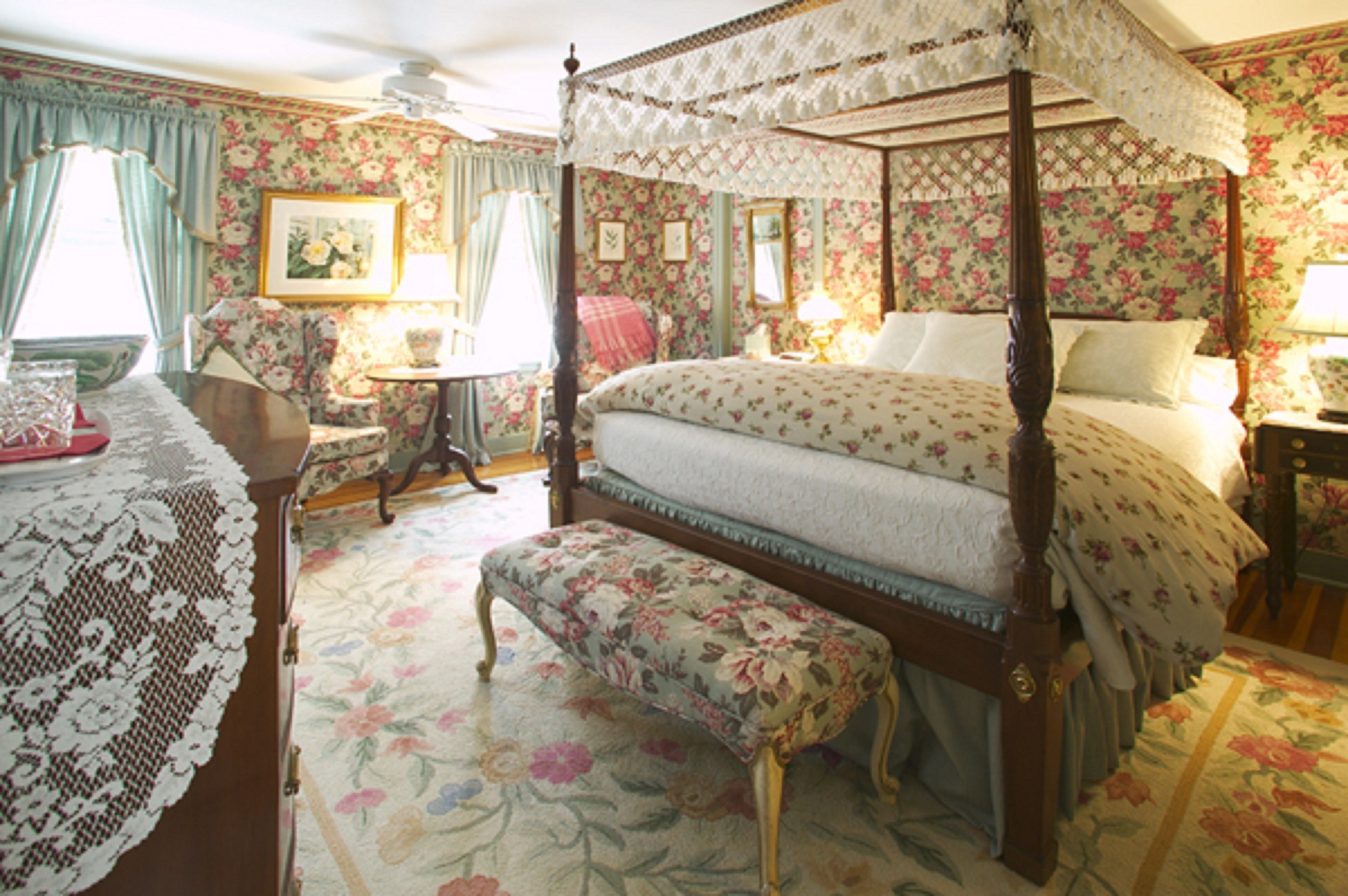 A four-poster canopy bed with floral upholstery, a matching bench, and a lace table runner.