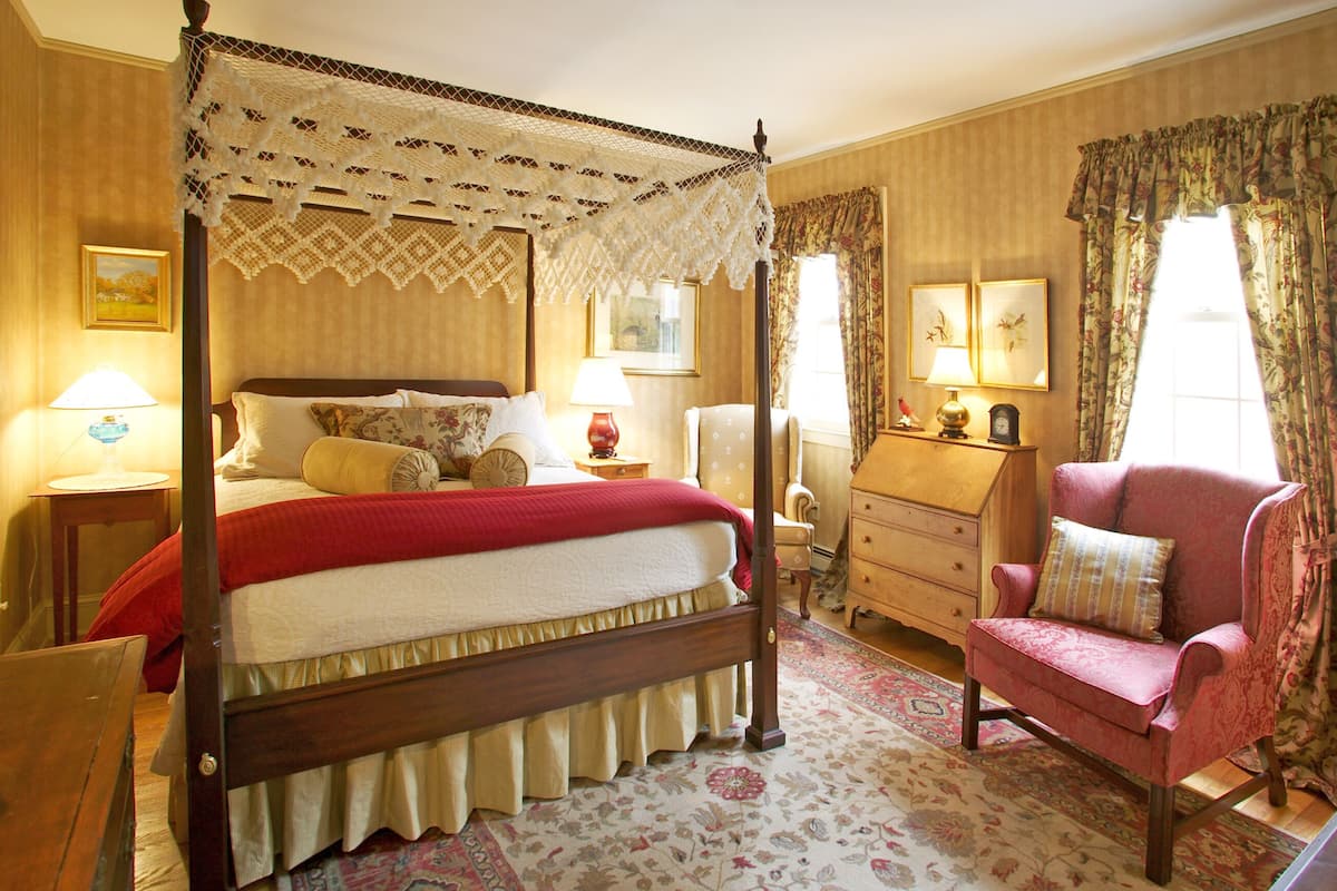 A traditional bedroom with a four-poster bed, a wooden dresser, a vanity with a mirror, and a red armchair.