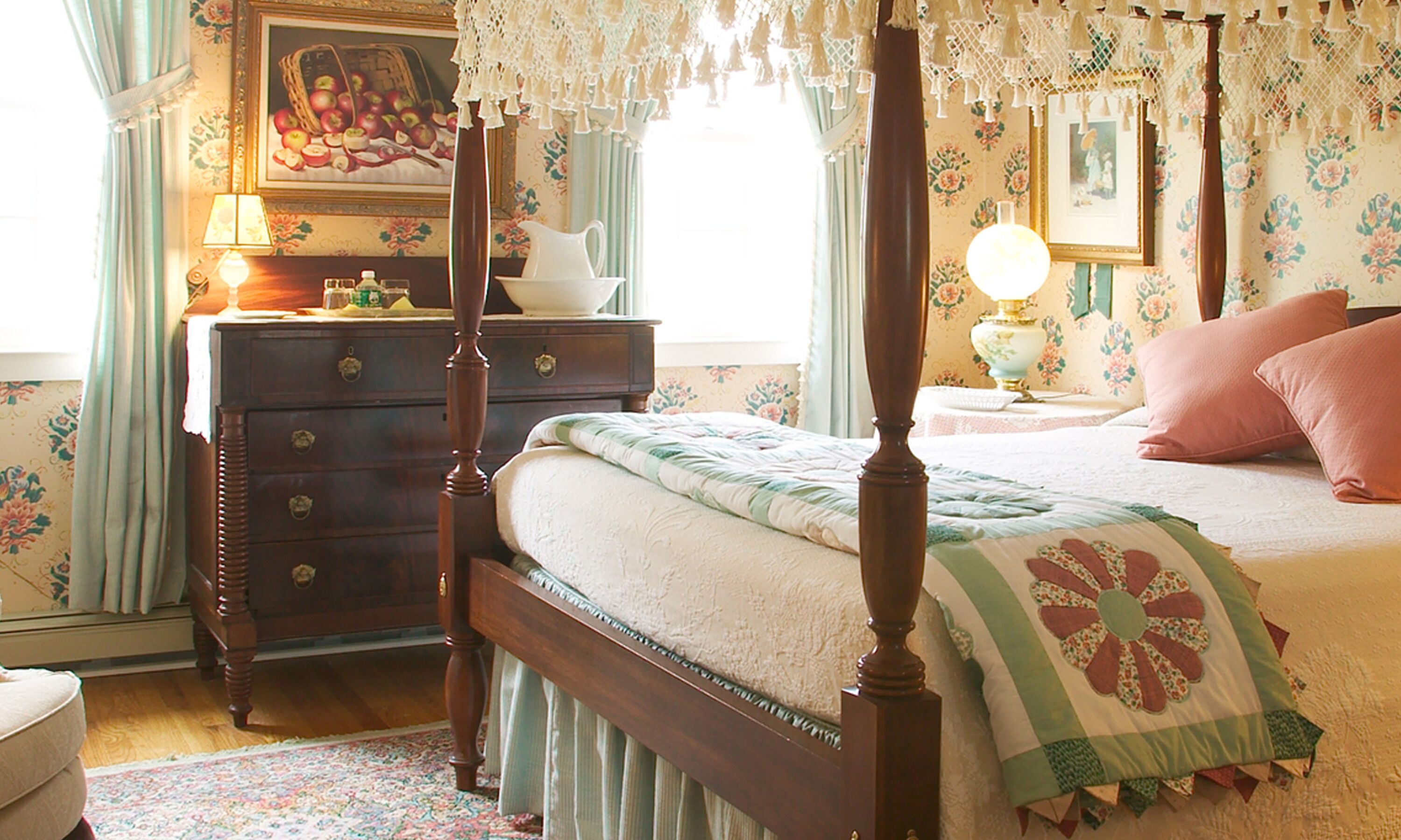 A four-poster bed with a floral bedspread, a nightstand with a lamp, a dresser with drawers, and a painting on the wall.