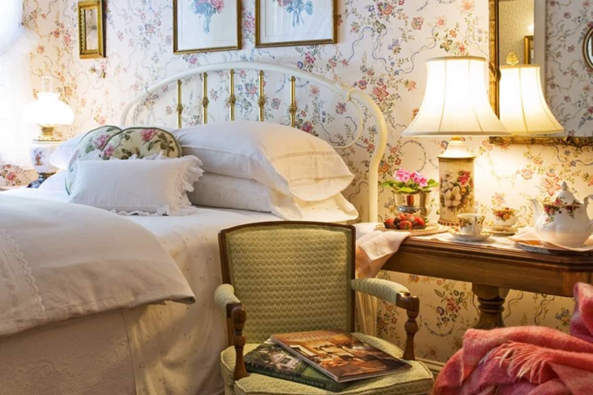 A bedroom with a bed, a chair, a bedside table with a lamp, and floral wallpaper.