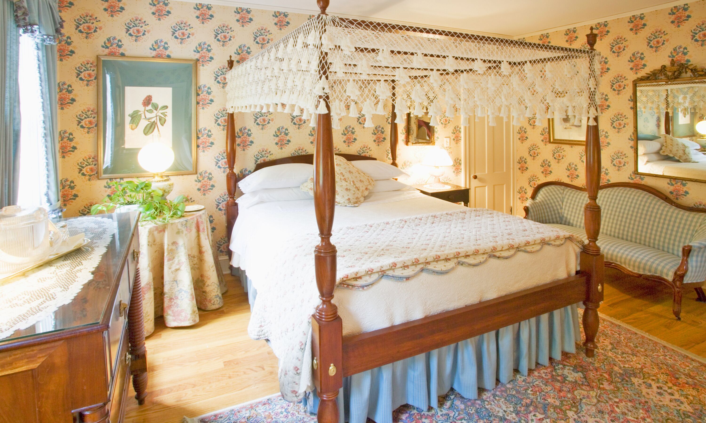 A bedroom with a four-poster bed, floral wallpaper, a round table with a lace tablecloth, and a chair with a checkered cushion.