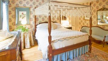 A bedroom with a four-poster bed, floral wallpaper, a round table with a lace tablecloth, and a chair with a checkered cushion.