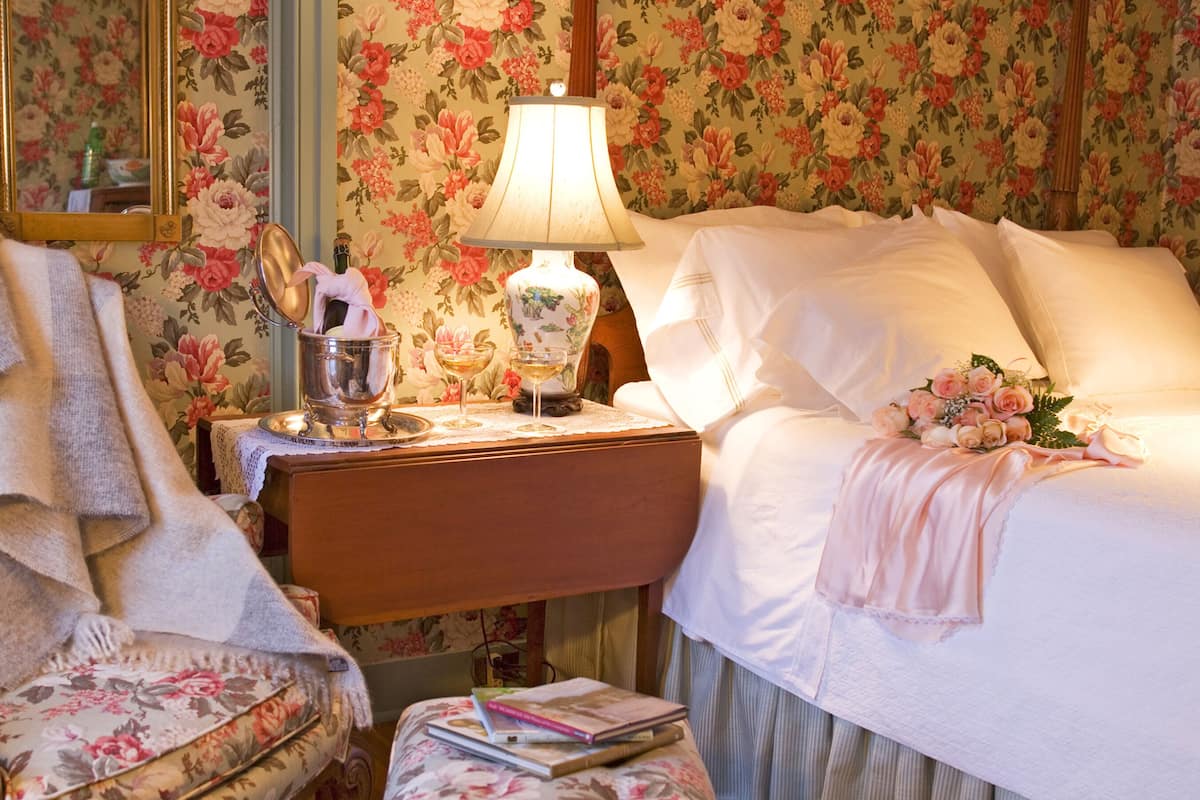 A bedroom with floral wallpaper, a bedside table with a lamp, a bed with a pink blanket, and a chair with a floral cushion.