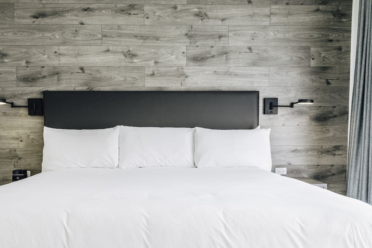Quarto moderno com parede de madeira, cabeceira preta, roupa de cama branca e duas luminárias de parede para leitura.