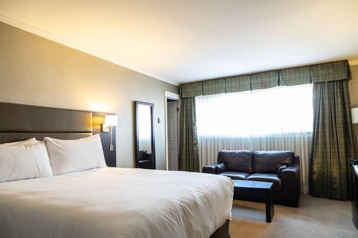 Quarto de hotel com uma cama grande, um sofá de couro, uma mesinha e uma janela com cortinas.