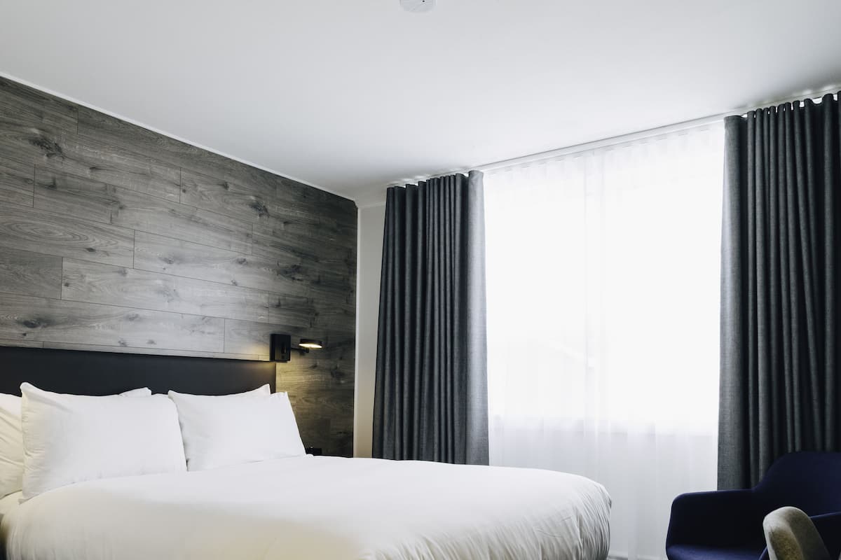 Quarto de hotel moderno com cabeceira de madeira, uma cama grande com lençóis brancos, uma poltrona azul escura e uma janela ampla com cortinas.