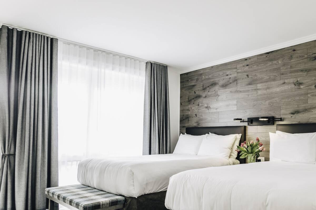 Quarto de hotel com duas camas, um banco, parede de destaque em madeira e janela com cortinas.
