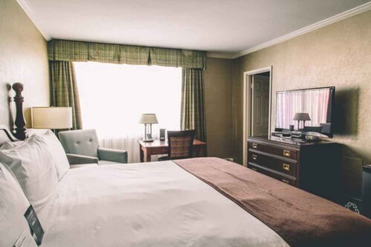 Quarto de hotel com uma cama grande, uma mesa com luminária, uma cadeira, cômoda e televisão.