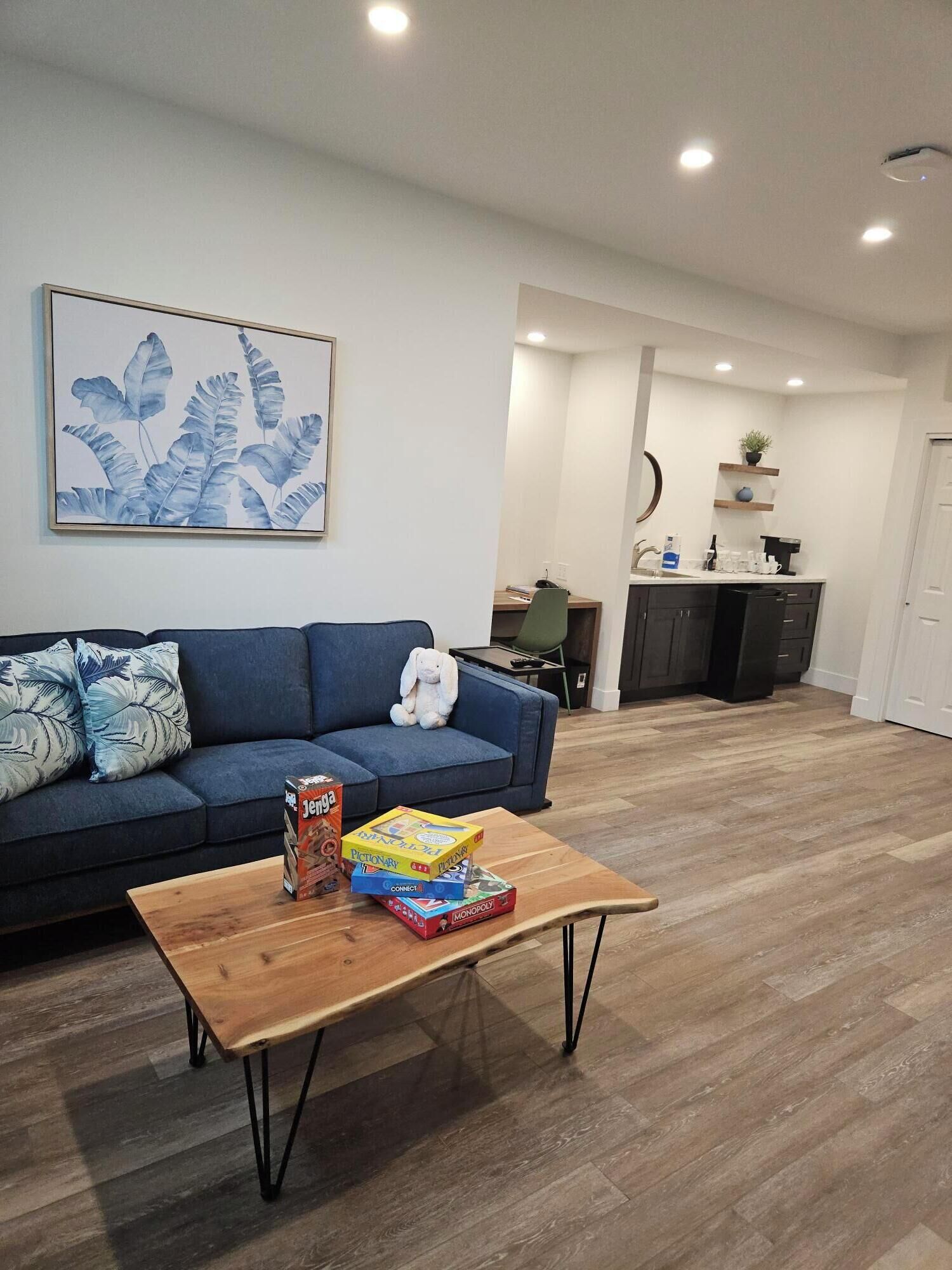 A living room with a sofa, a coffee table with board games, a painting on the wall, and a kitchen area in the background.