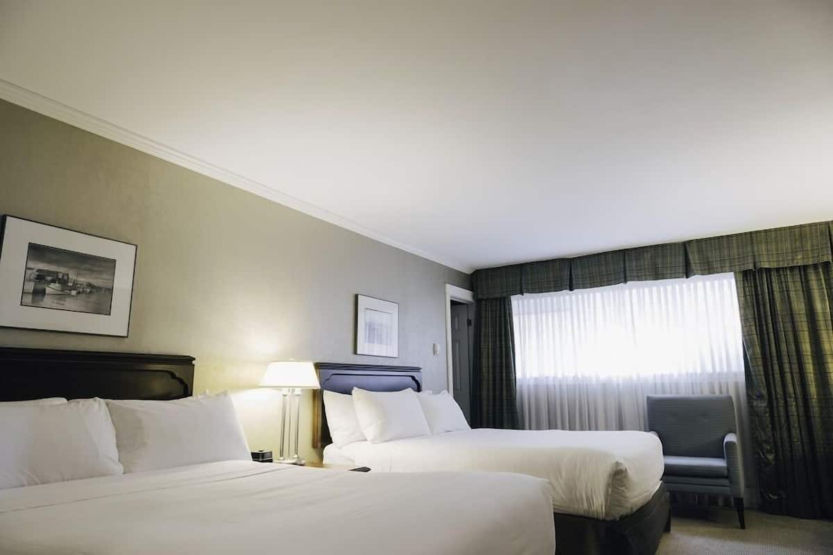 Quarto de hotel com duas camas, uma cadeira, uma luminária e uma janela com cortinas.