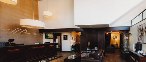 Um hall de hotel com uma recepção, uma área de estar com sofá e mesa de centro, e uma escadaria que leva a um andar superior.