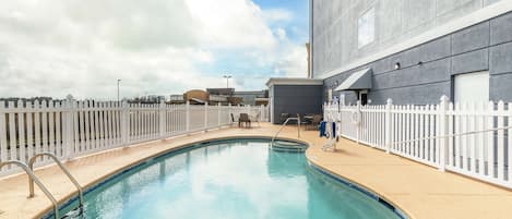 Una piscina con cerco blanco, un edificio de fachada gris y un cielo despejado.