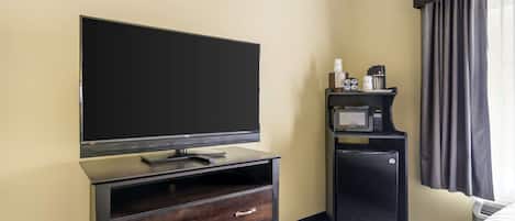 Habitación de hotel con televisión de pantalla plana sobre un soporte de madera, un refrigerador pequeño, un microondas y una cama con colcha estampada.