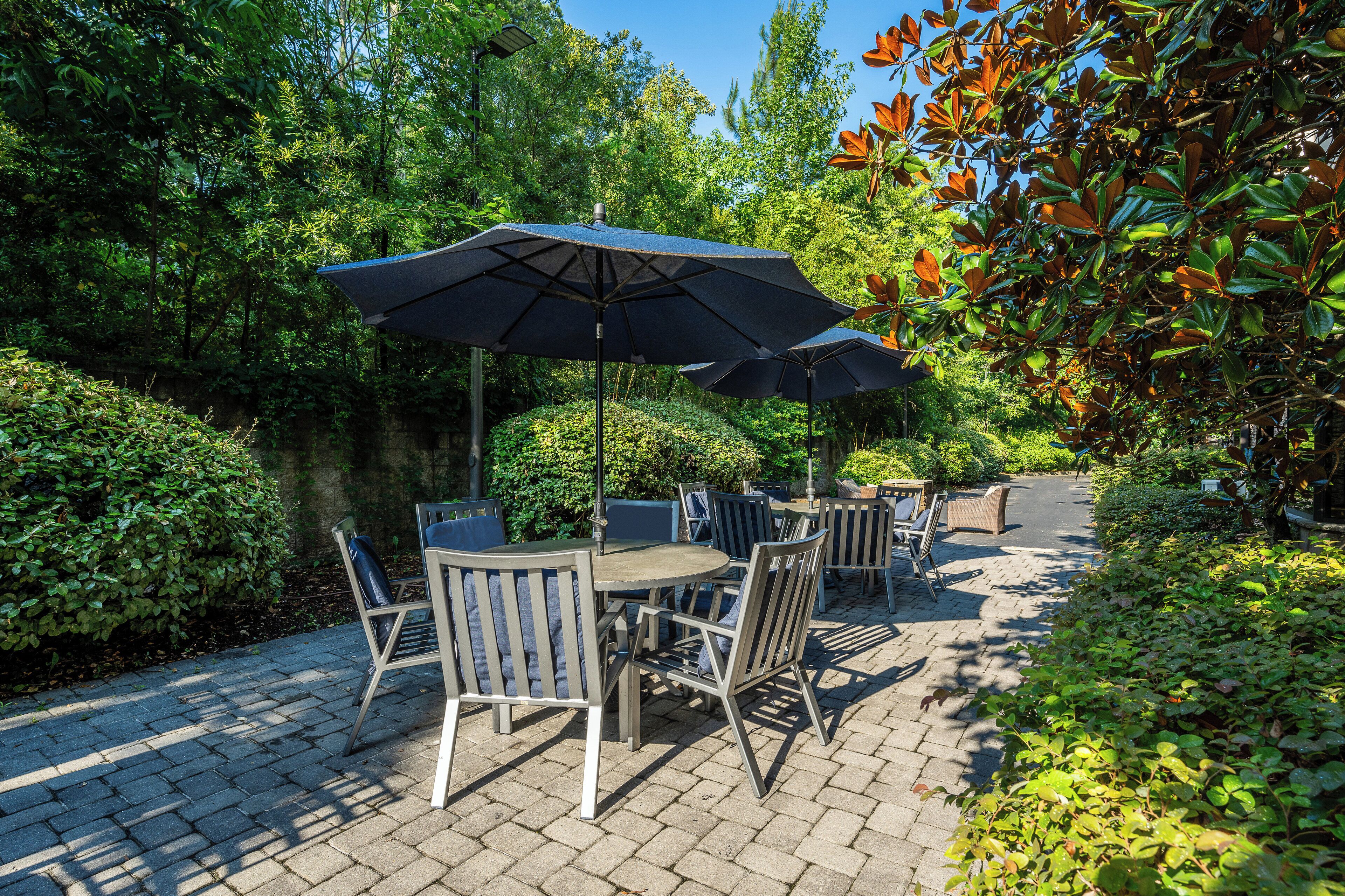 Eine Terrasse im Freien mit Tischen und Stühlen, umgeben von üppiger Bepflanzung und Bäumen.