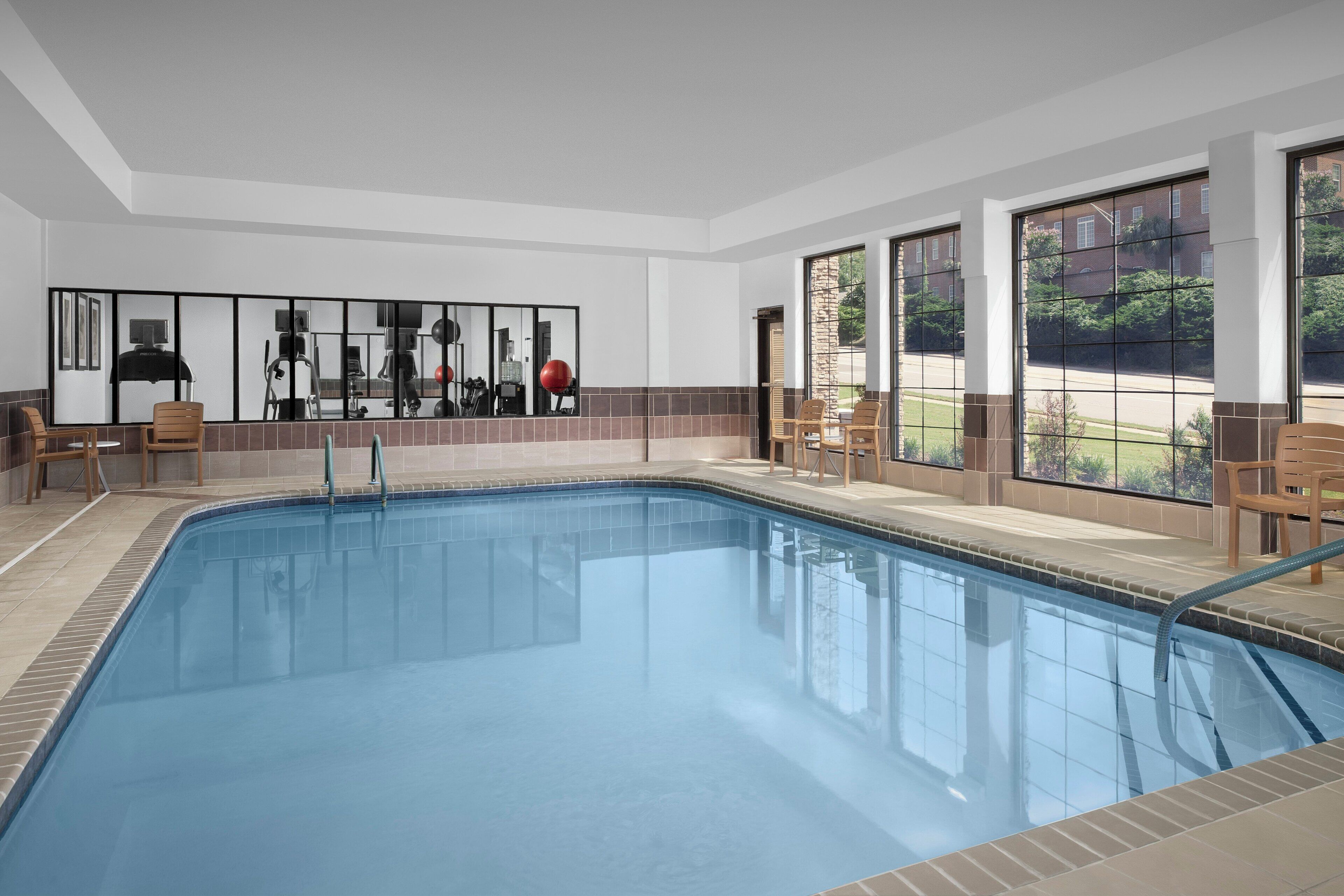 A swimming pool with a tiled deck, exercise equipment, and large windows offering views of the outdoors.