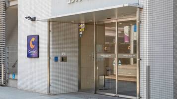 Entrada de un hotel con cartel de Comfort Hotel, símbolo de AED y señal de accesibilidad para sillas de ruedas.