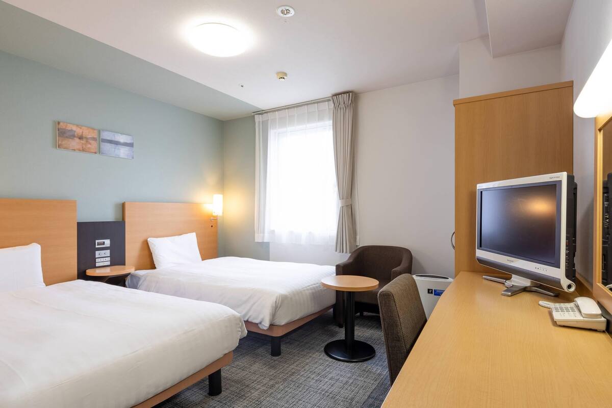 A hotel room with two beds, a desk with a TV, a chair, a small table, and a window with curtains.