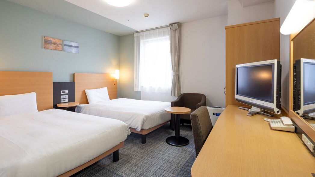 A hotel room with two beds, a desk with a computer and phone, a chair, and a window with curtains.