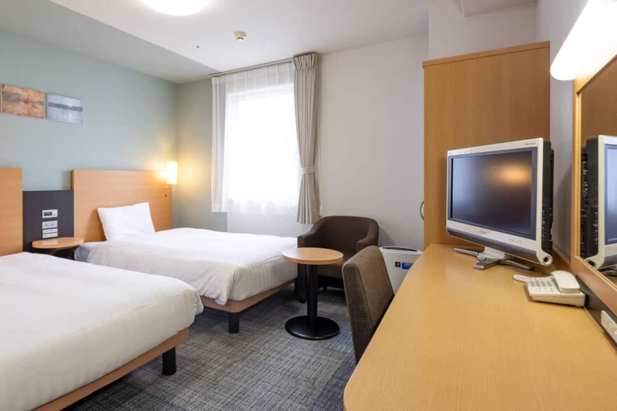 Ein Hotelzimmer mit zwei Betten, einem Schreibtisch mit Computer und Telefon, einem Sessel und einem Fenster mit Vorhängen.