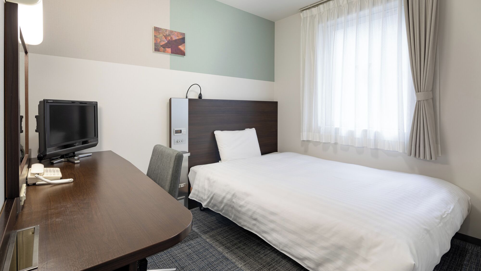 A hotel room with a bed, a desk with a computer, a chair, a bedside table, a lamp, and a window with curtains.