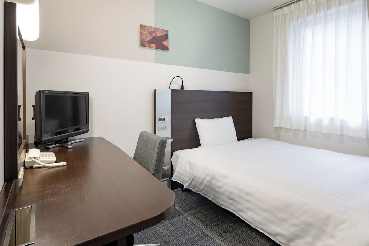 Ein Hotelzimmer mit Bett, Schreibtisch mit Computer, Stuhl, Nachttisch, Lampe und einem Fenster mit Vorhängen.