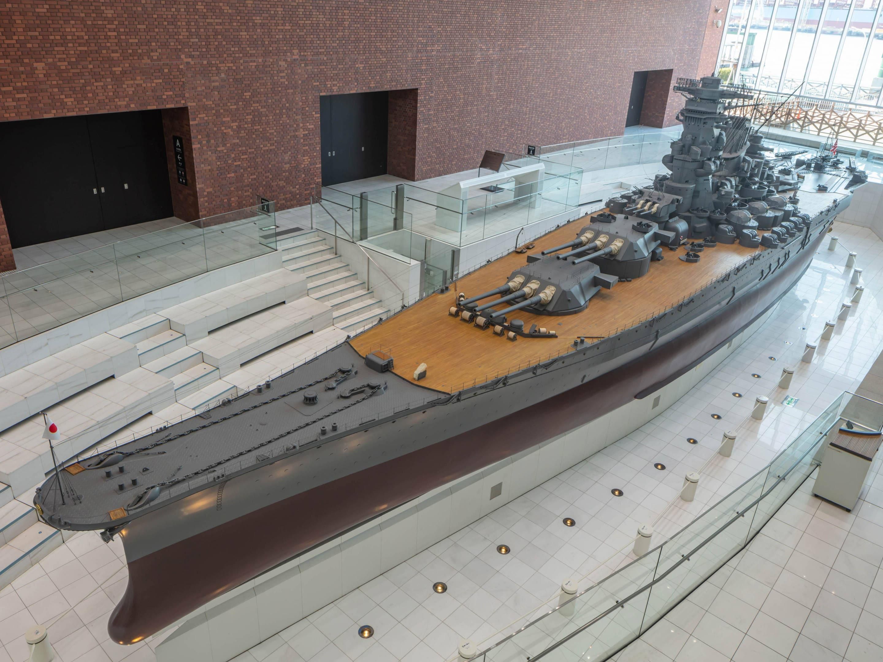 A large ship model displayed indoors with glass railings, a wooden deck, and a brick wall in the background.