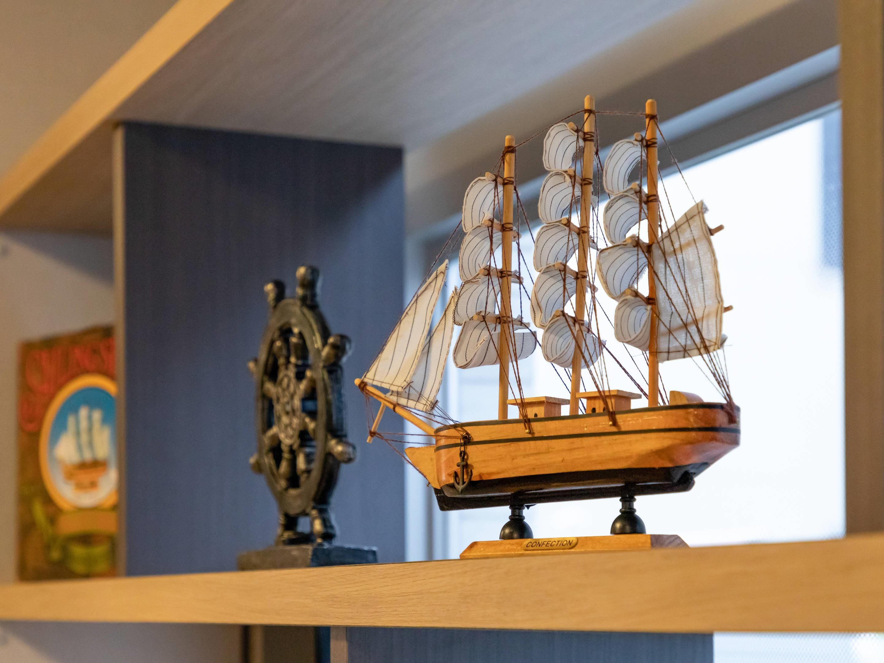A model ship on a shelf with a maritime-themed painting and a ship's wheel in the background.