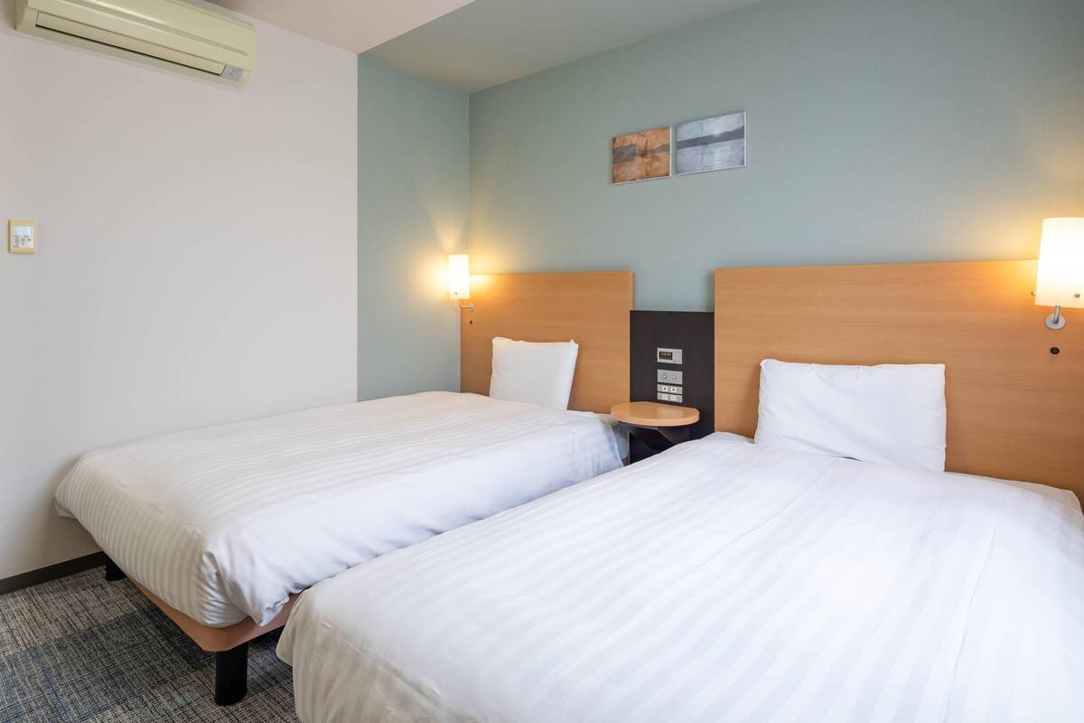 Ein Hotelzimmer mit zwei Einzelbetten, einem kleinen Nachttisch, einer Wand-Klimaanlage und einem Beistelltischchen mit Lampe.