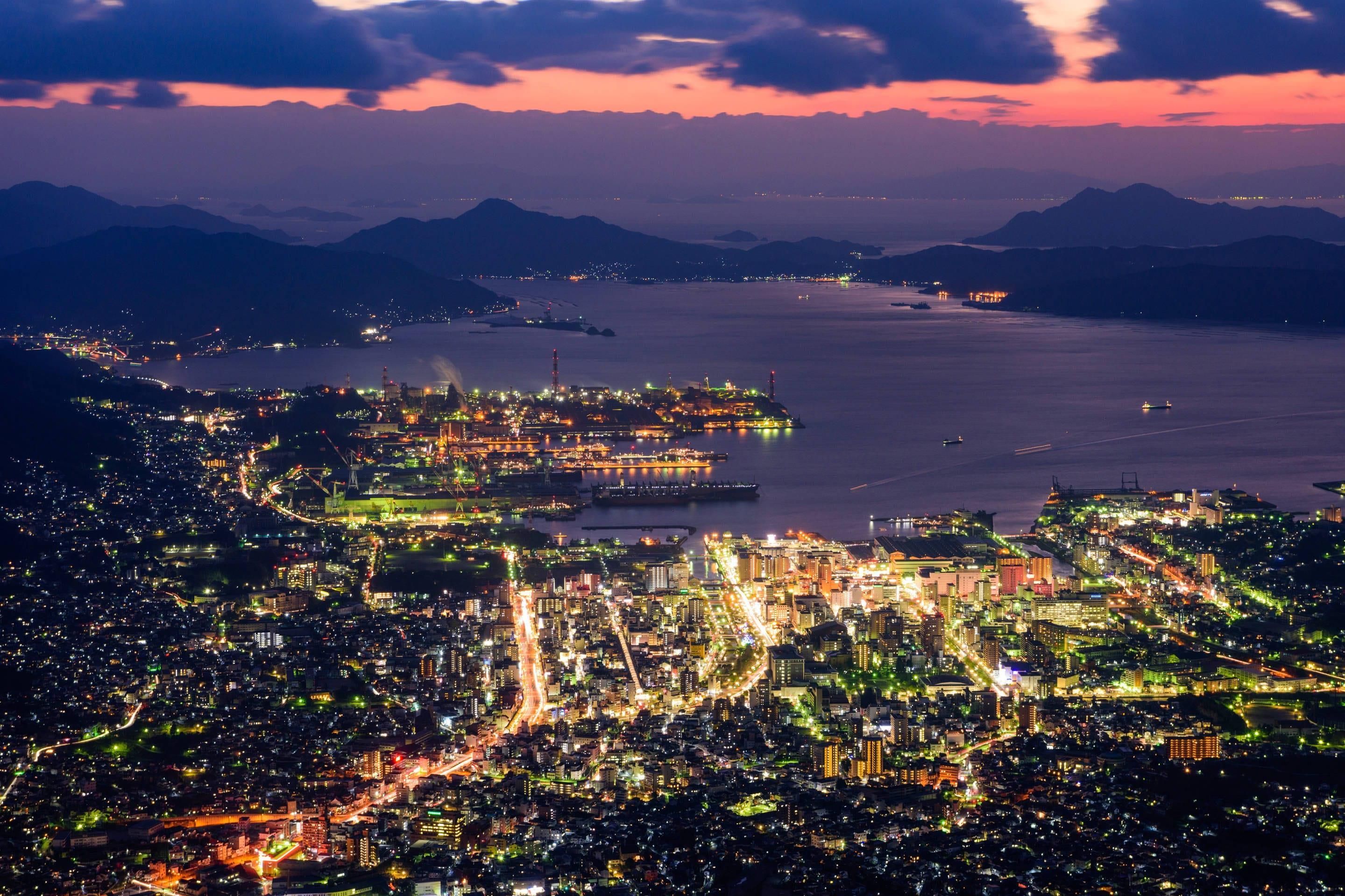 A nighttime aerial view of a coastal city with illuminated buildings and a harbor.