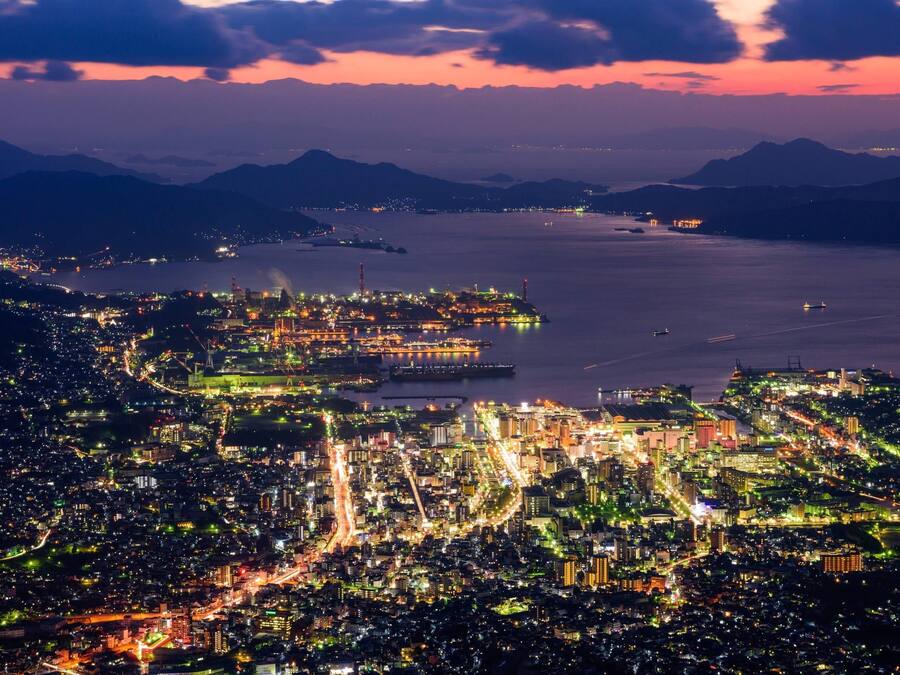 A nighttime aerial view of a coastal city with illuminated buildings and a harbor.