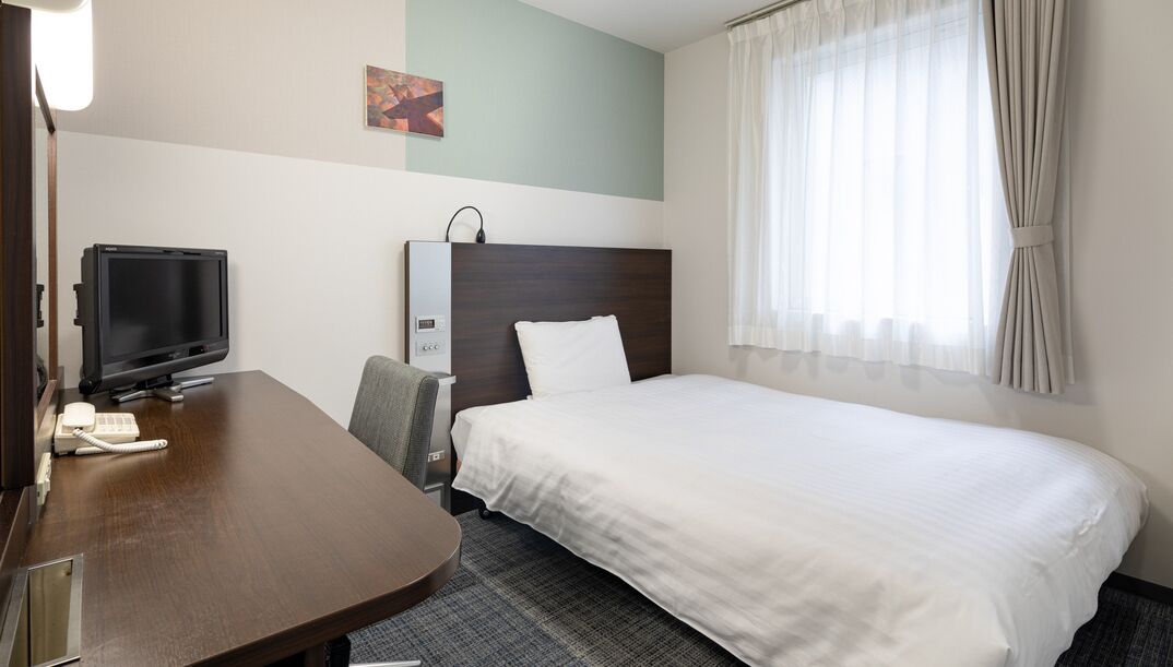 A hotel room with a bed, a desk with a television, a chair, and a window with curtains.