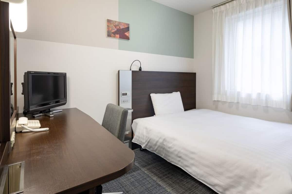 Ein Hotelzimmer mit Bett, Schreibtisch, Fernseher, Stuhl und einem Fenster mit Vorhängen.