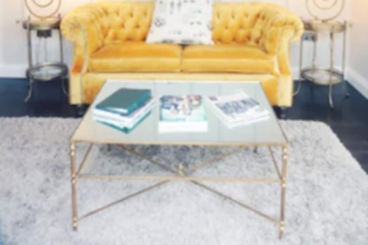 Un sofá amarillo con diseño capitonado, una mesa de centro de vidrio y dos lámparas sobre mesitas.