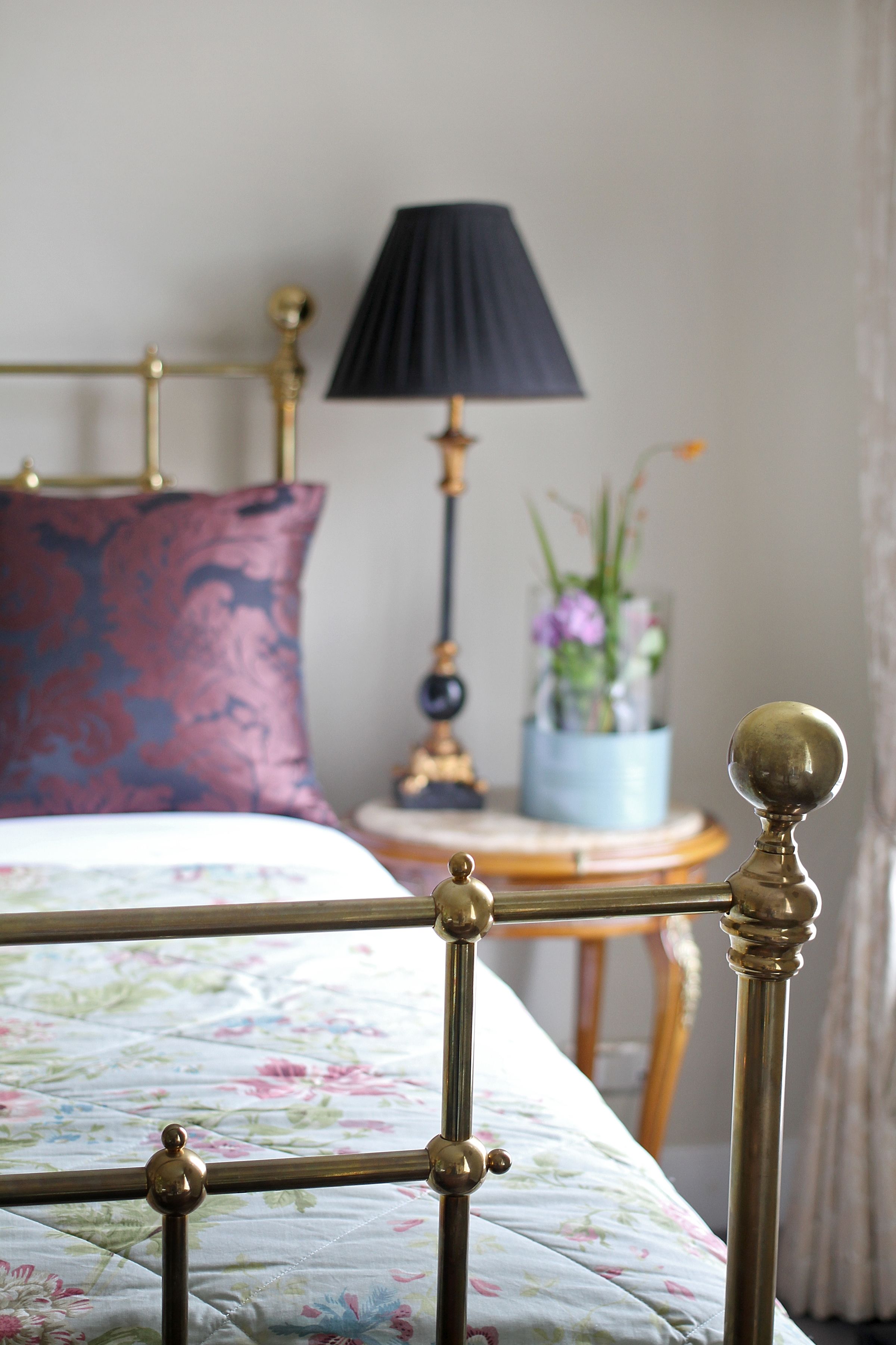 A bed with a floral bedspread, a brass headboard, a dark blue lamp, and a small table with a vase of flowers.