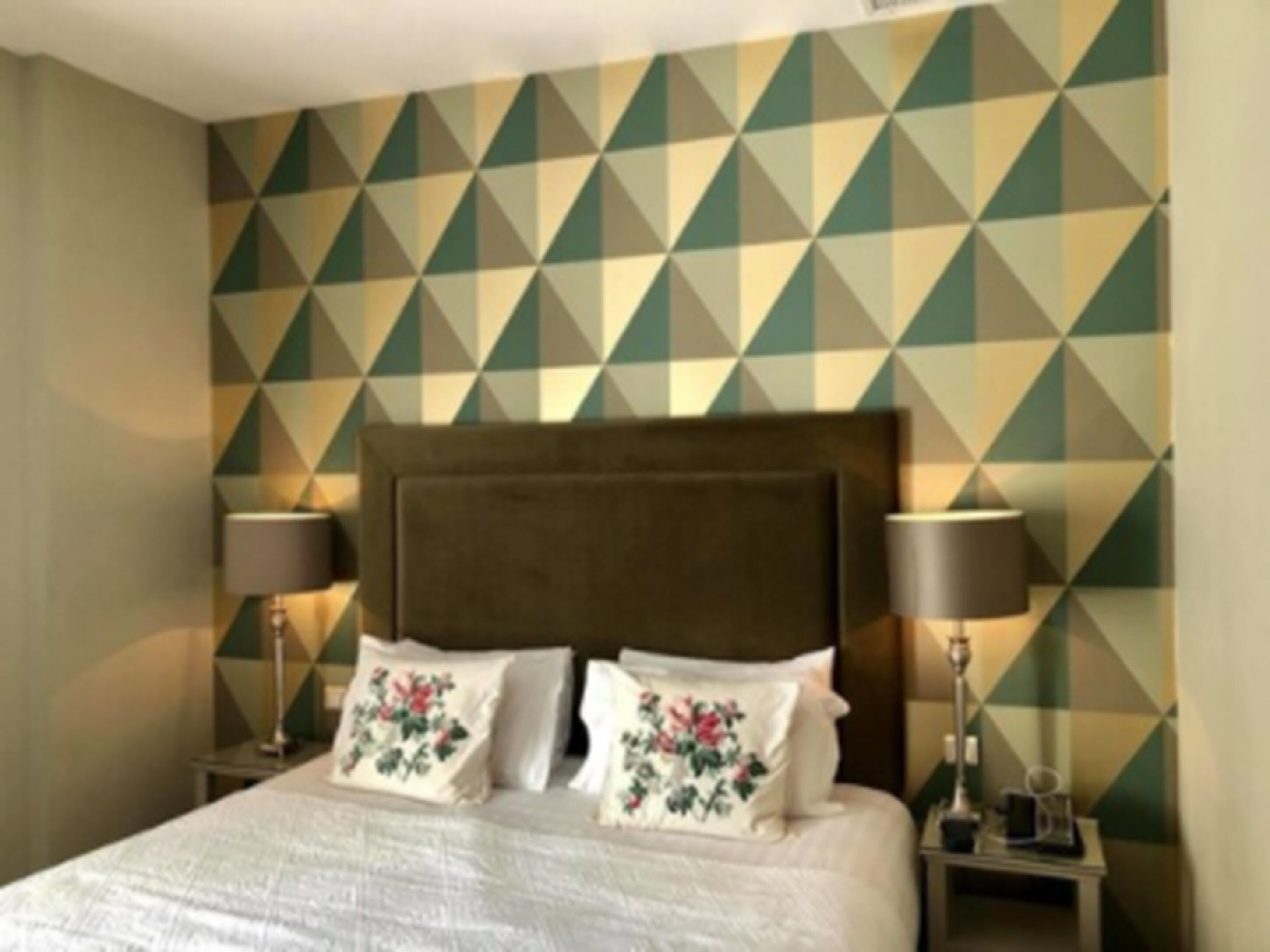 Habitación de hotel con pared de diseño geométrico, una cama grande con almohadas florales y dos lámparas de noche.
