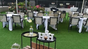 Un espacio para eventos al aire libre con mesas redondas, manteles blancos y sillas negras.