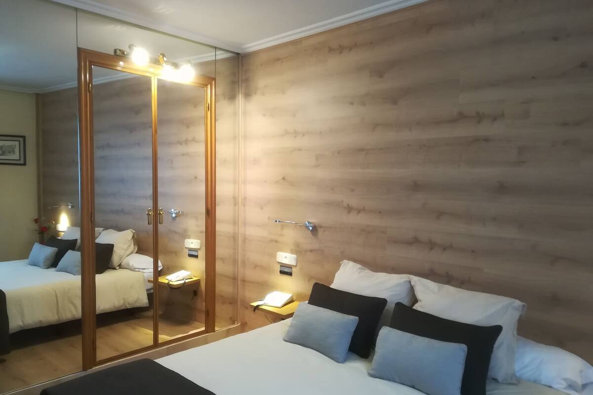 Une chambre avec un grand miroir, des lambris en bois sur les murs, un lit avec une literie blanche et plusieurs oreillers, des tables de chevet avec des lampes et une petite table de nuit avec un téléphone.