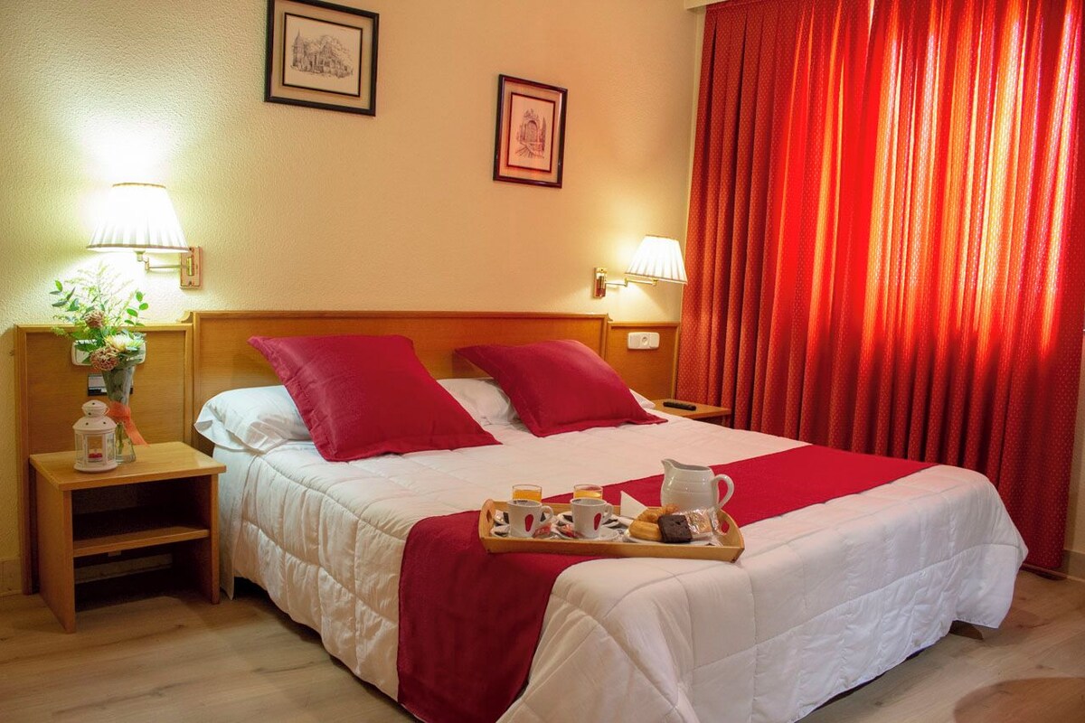 Ein Hotelzimmer mit einem Bett, zwei Kissen, einem Tablett mit Frühstücksangeboten, einem Nachttisch mit Lampe und einem Fenster mit roten Vorhängen.