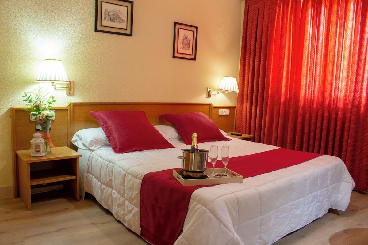 Ein Hotelzimmer mit einem großen Bett, roten Kissen, einem Tablett mit einer Flasche und Gläsern sowie zwei Nachttischlampen.