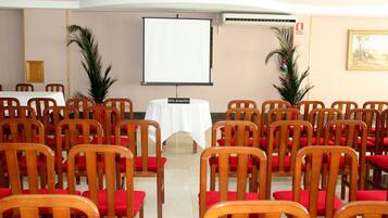 Un salón de conferencias con filas de sillas de madera con cojines rojos, una mesa blanca con un cartel y una pantalla de proyector en la pared.