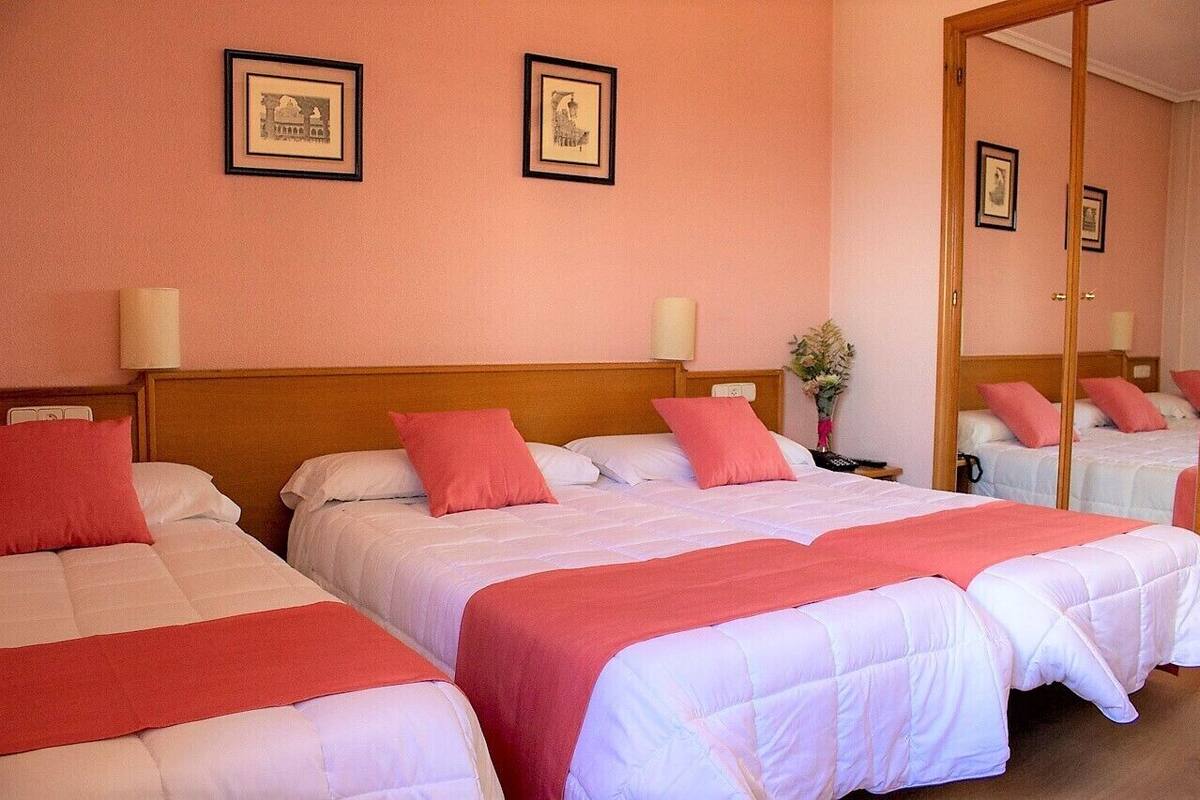 Une chambre d’hôtel avec deux lits, chacun doté d’une tête de lit en bois, de draps blancs et d’éléments décoratifs rouges. Des tableaux encadrés ornent le mur, et un miroir reflète l’intérieur de la pièce.