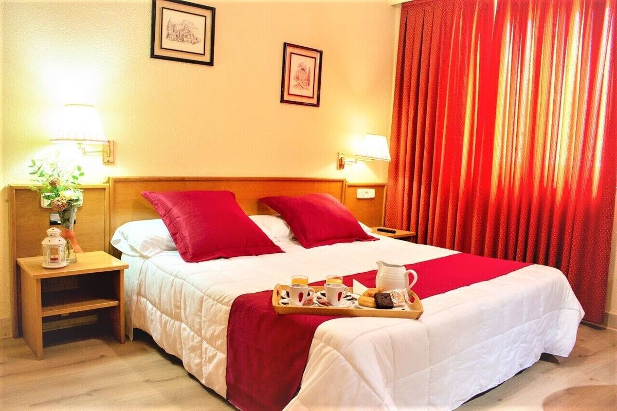 Une chambre d’hôtel avec un lit, des rideaux rouges, un plateau contenant des articles pour le petit-déjeuner et deux lampes fixées au mur.