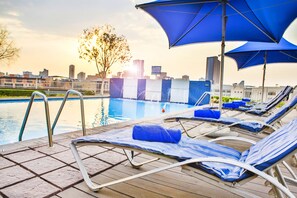 Un espace aménagé au bord de la piscine, avec des chaises longues et des parasols, une vue dégagée sur les gratte-ciel de la ville et un arbre en arrière-plan.