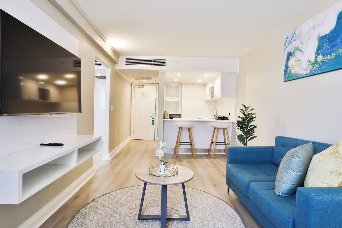 Una sala de estar moderna con una televisión de pantalla plana, un sofá azul, una mesita redonda y una cocina al fondo.